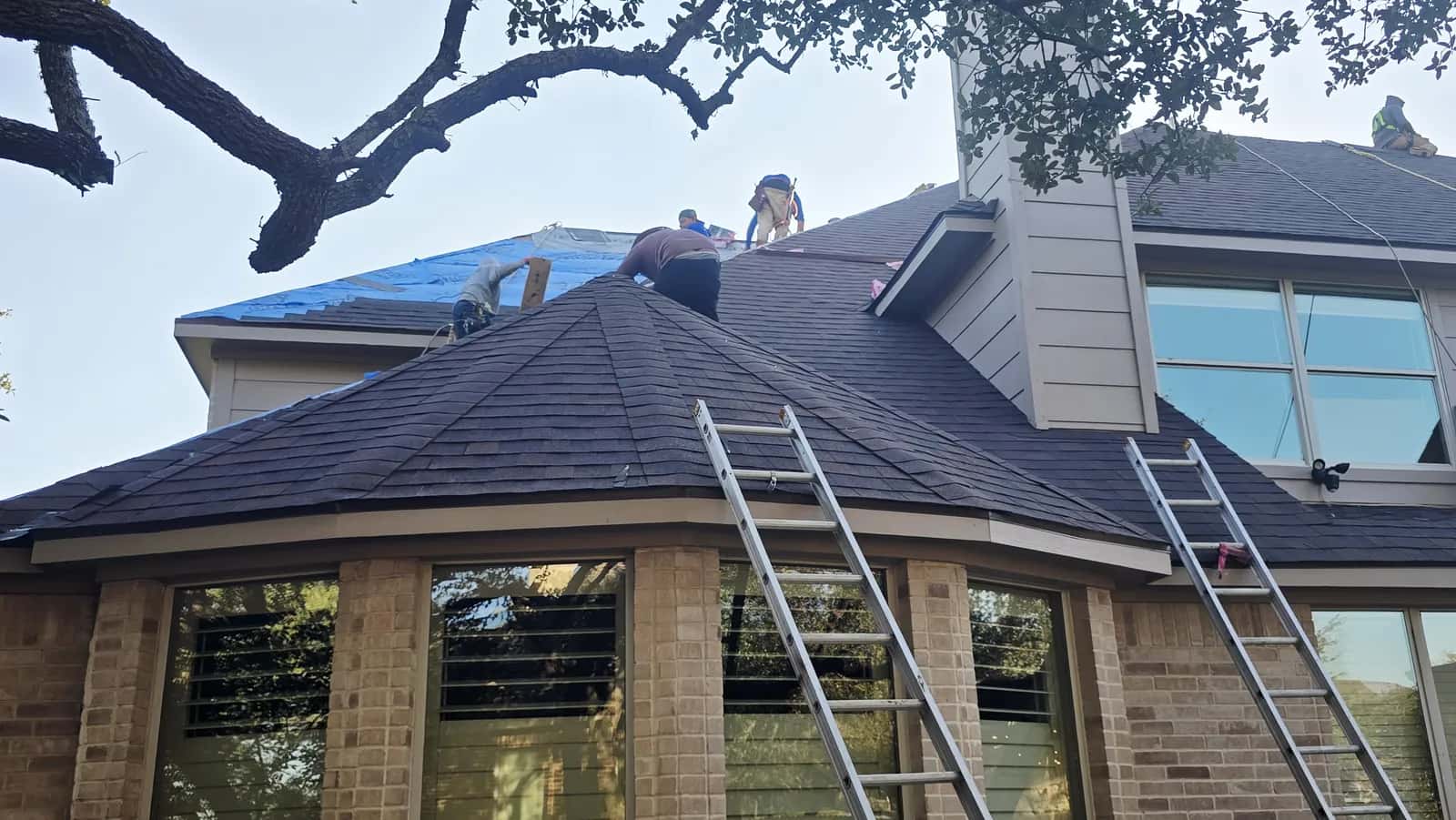 Crew installing dark gray architectural shingles over blue synthetic underlayment above a brick bay window, ladders at the eave.