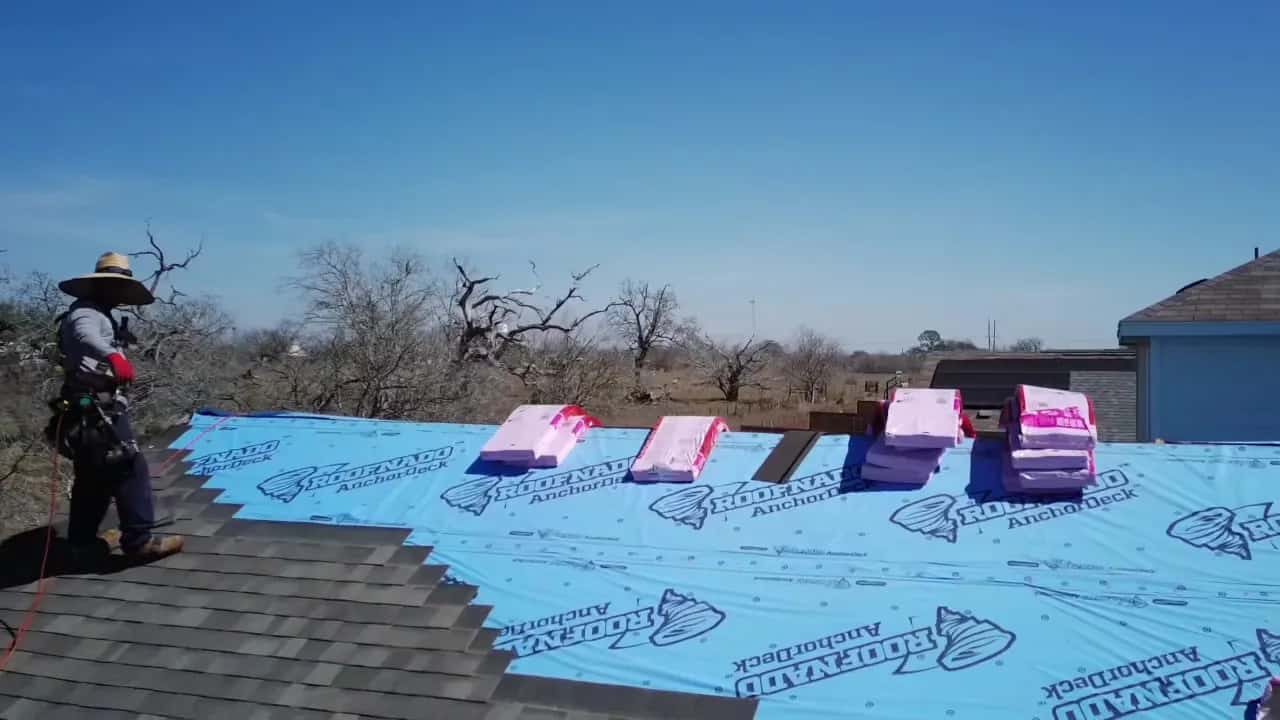 Roofer on blue synthetic underlayment with shingle bundles staged on the roof, wide open sky beyond.