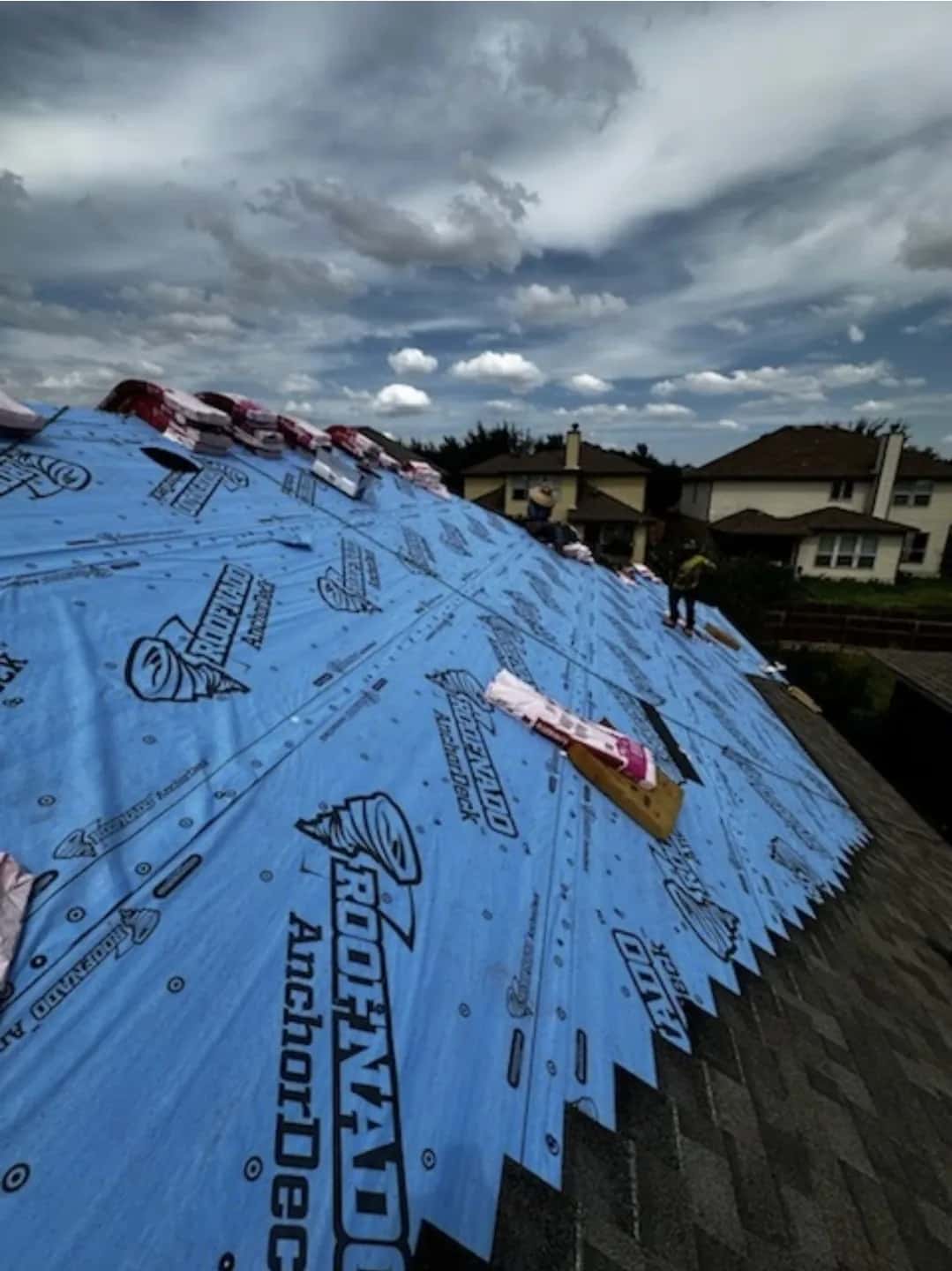 Blue synthetic underlayment with shingle bundles and crew at the ridge under a cloudy sky.