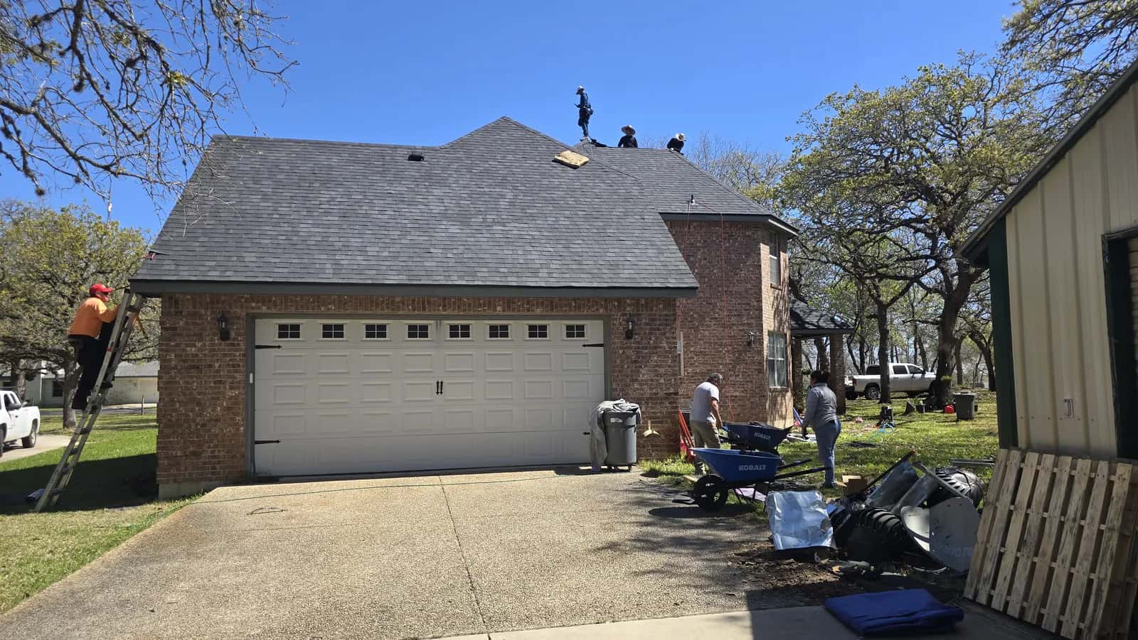 Brick home with a new gray shingle roof over the garage, crew cleaning up pallets and debris in the driveway.