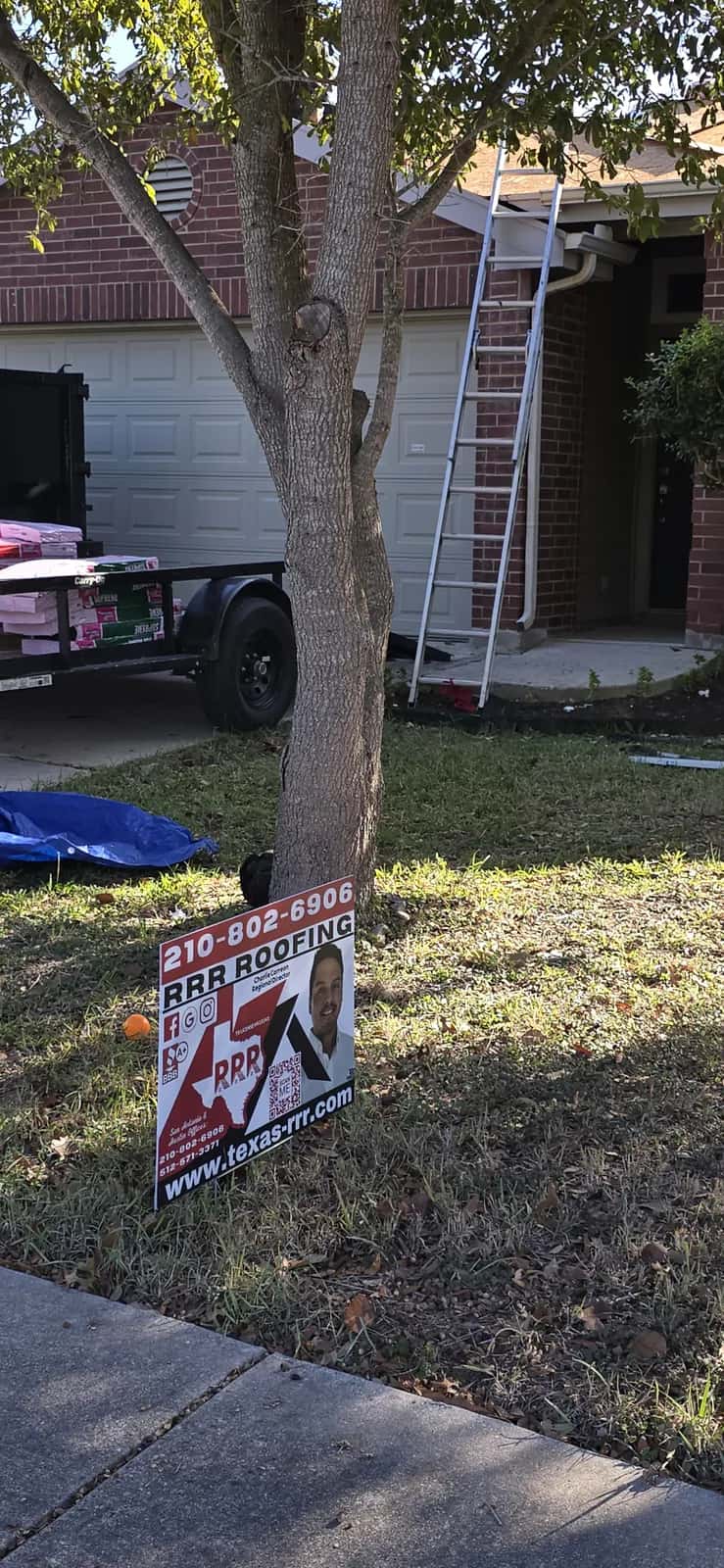 Front of a brick single-story home with an extension ladder on the roof, a trailer loaded with shingle bundles in the driveway, and an RRR Roofing yard sign on the lawn.