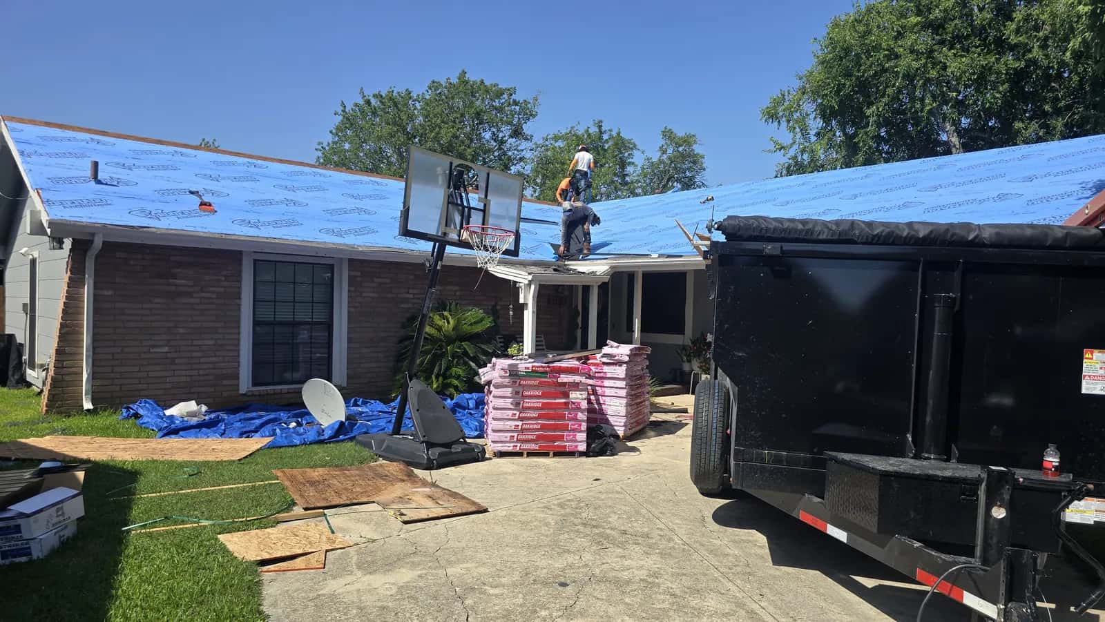 Brick ranch home with blue synthetic underlayment installed over the roof deck, shingle bundles staged on the driveway next to a dump trailer.