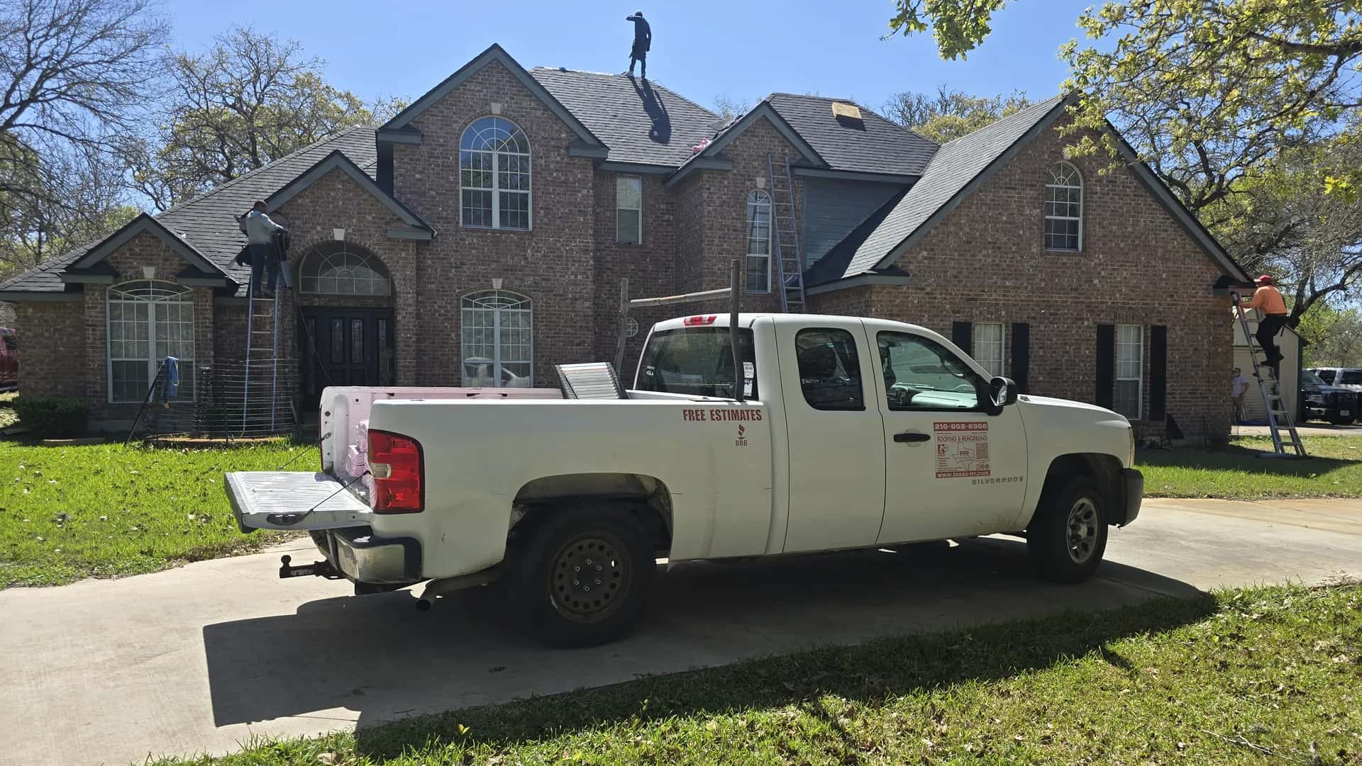 Two-story brick home with Texas-RRR roofers on the roof and ladders at the eaves, company pickup parked in the driveway.