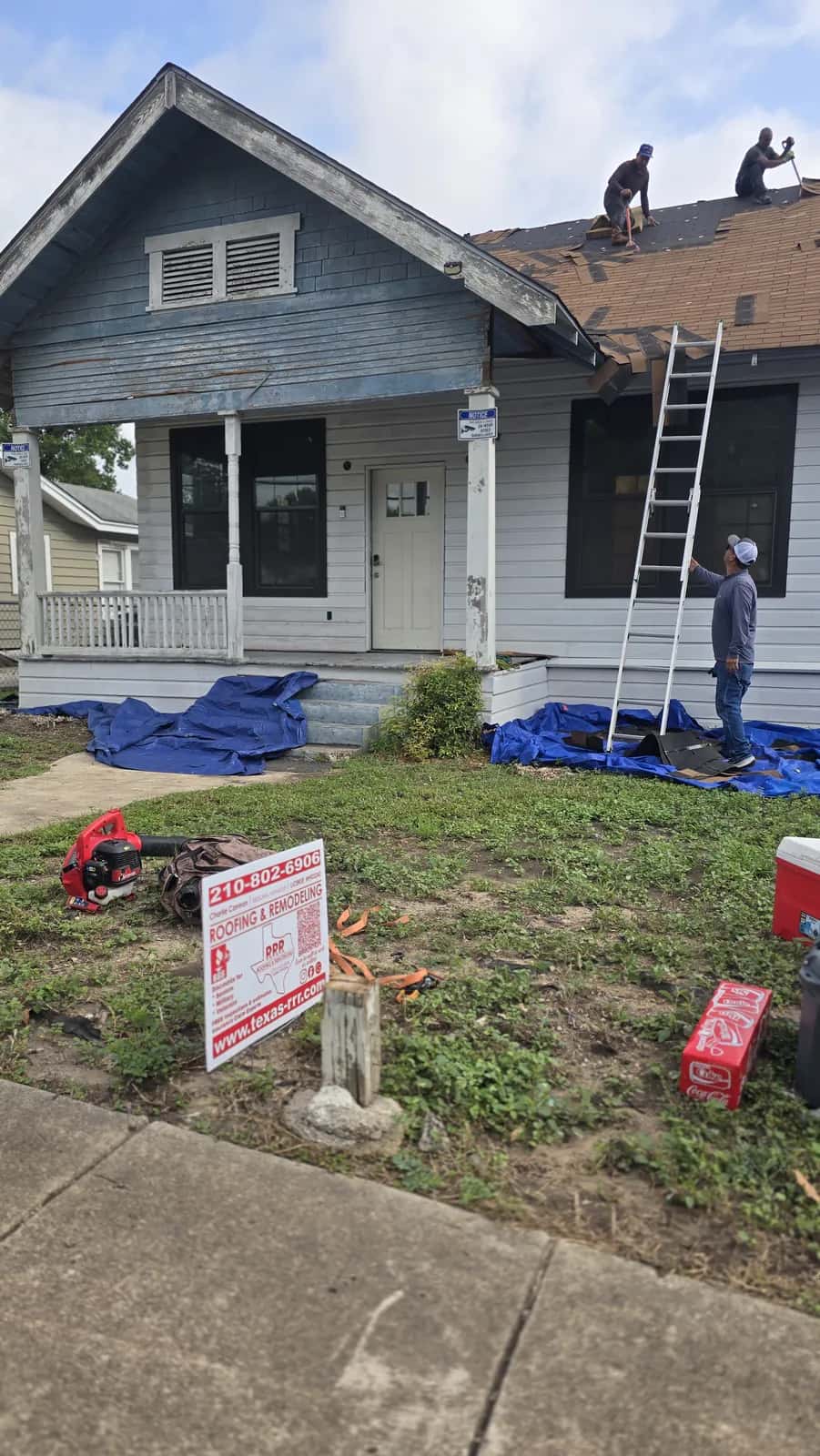 Crew tearing off old shingles on a wood-sided bungalow with weathered gable trim, tarps protecting the yard and a Texas-RRR sign out front