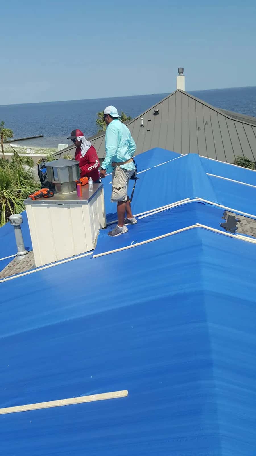 Two roofers on a roof covered in blue underlayment at a waterfront home, with a metal-roofed building in the background