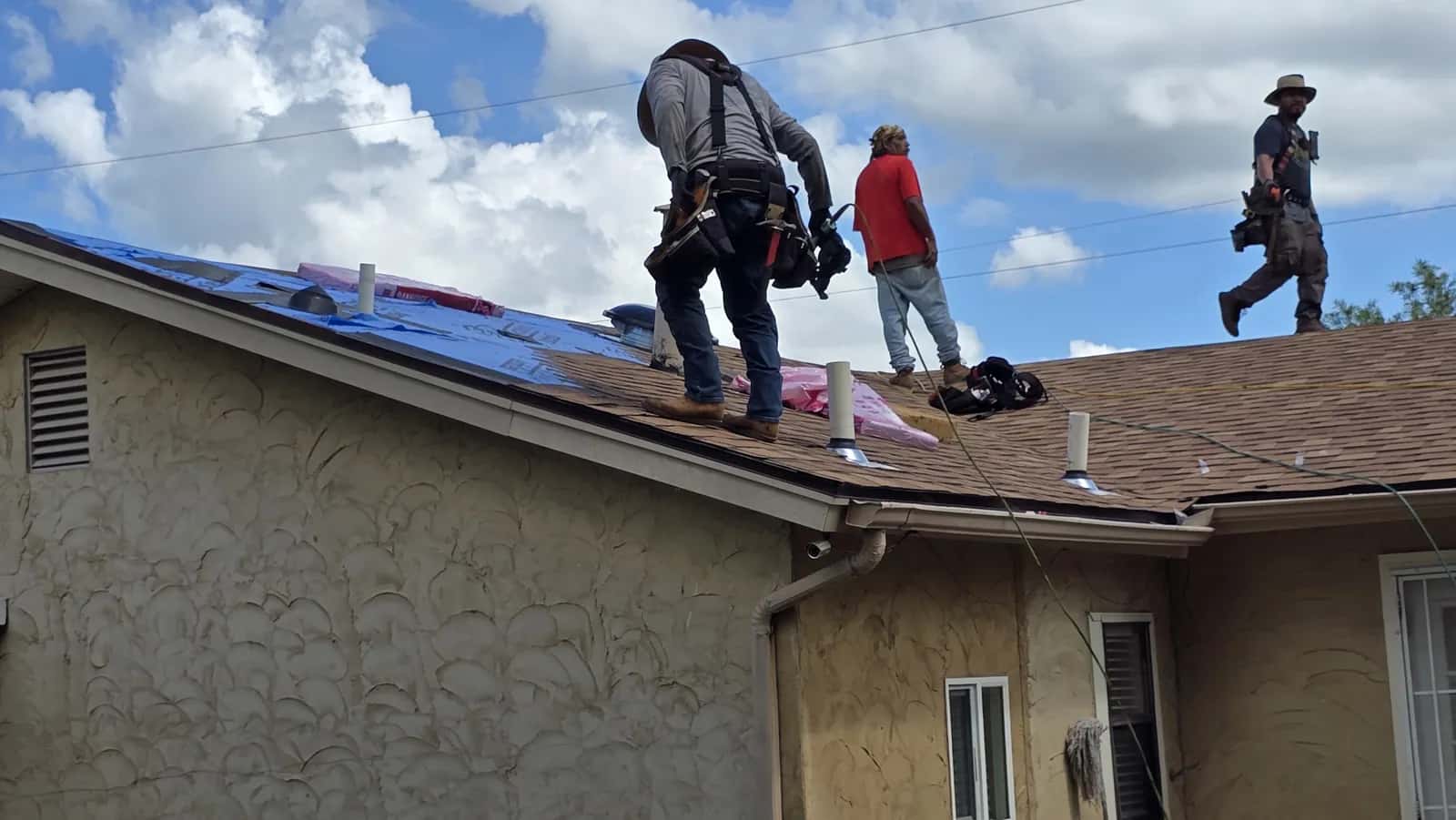 Three roofers installing brown architectural shingles over blue underlayment on a stucco home under a cloudy sky