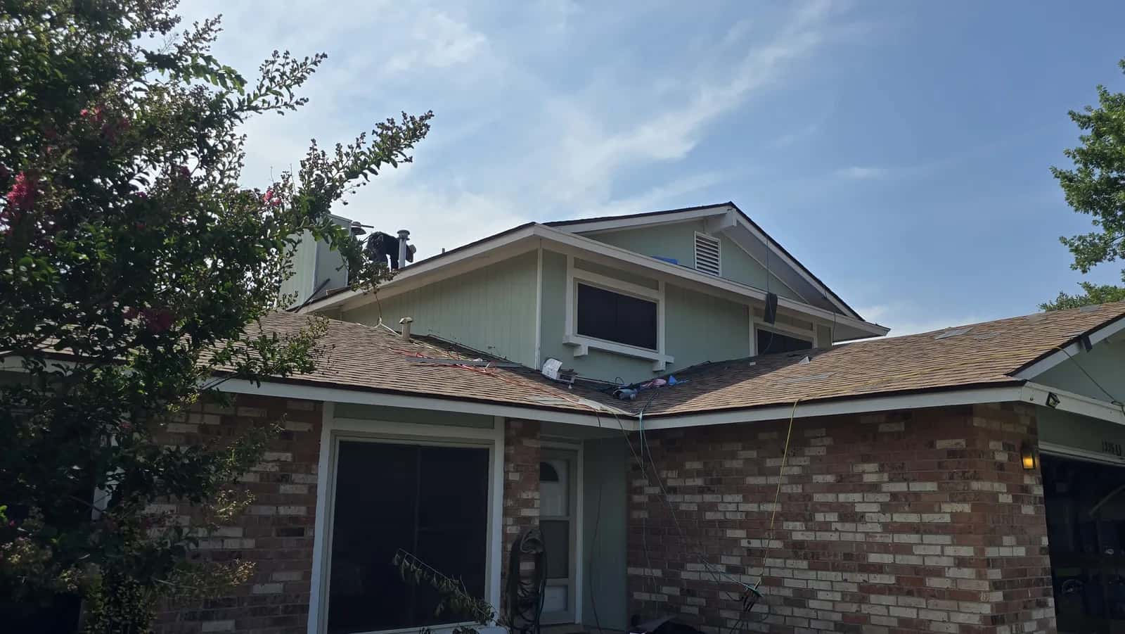 Single-story brick and siding home with a roofing crew at the ridge during shingle tear-off, old roofing removed down to the deck.