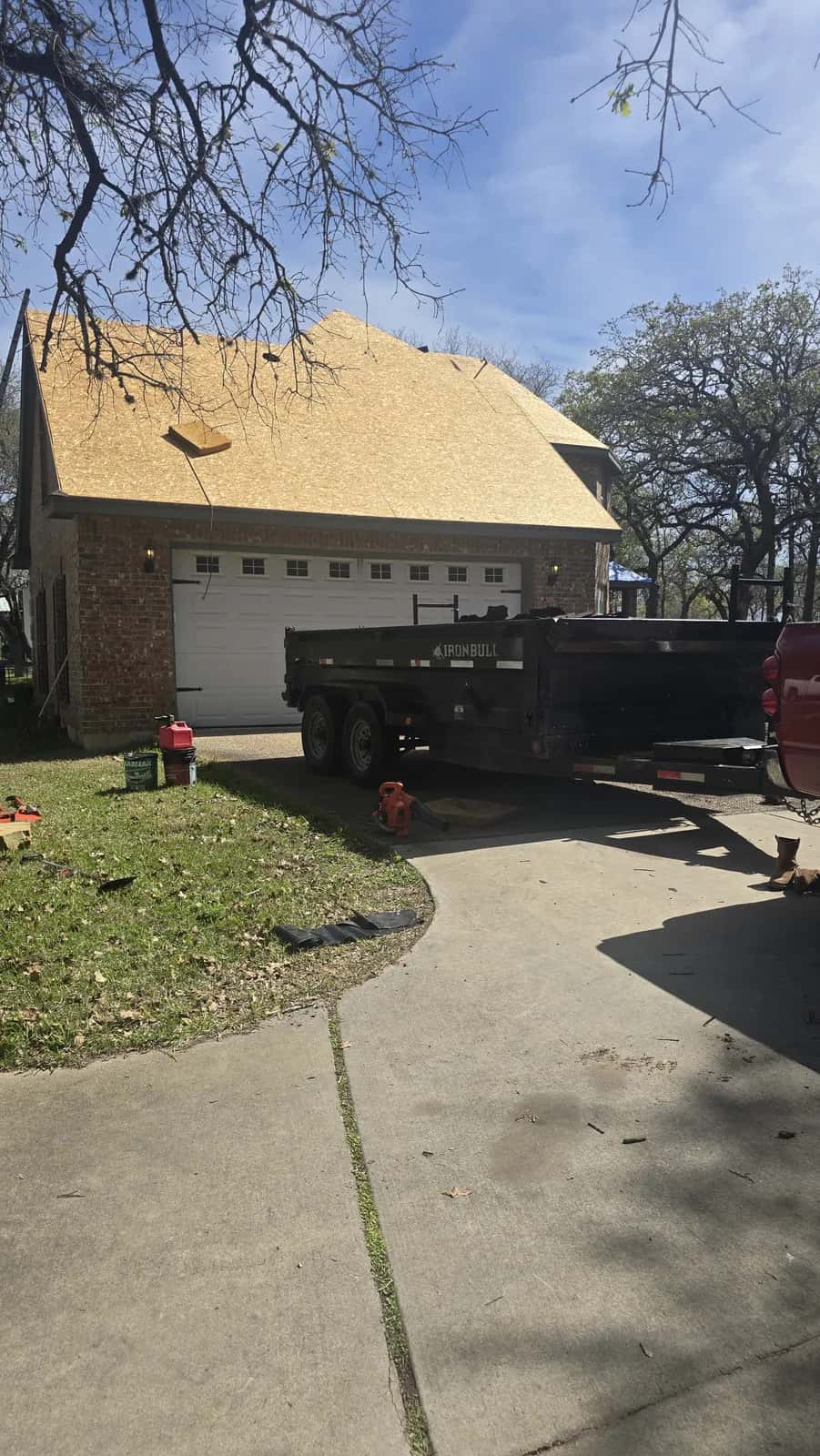 Two-story brick home with fresh OSB roof decking installed before shingles, dump trailer parked in the driveway