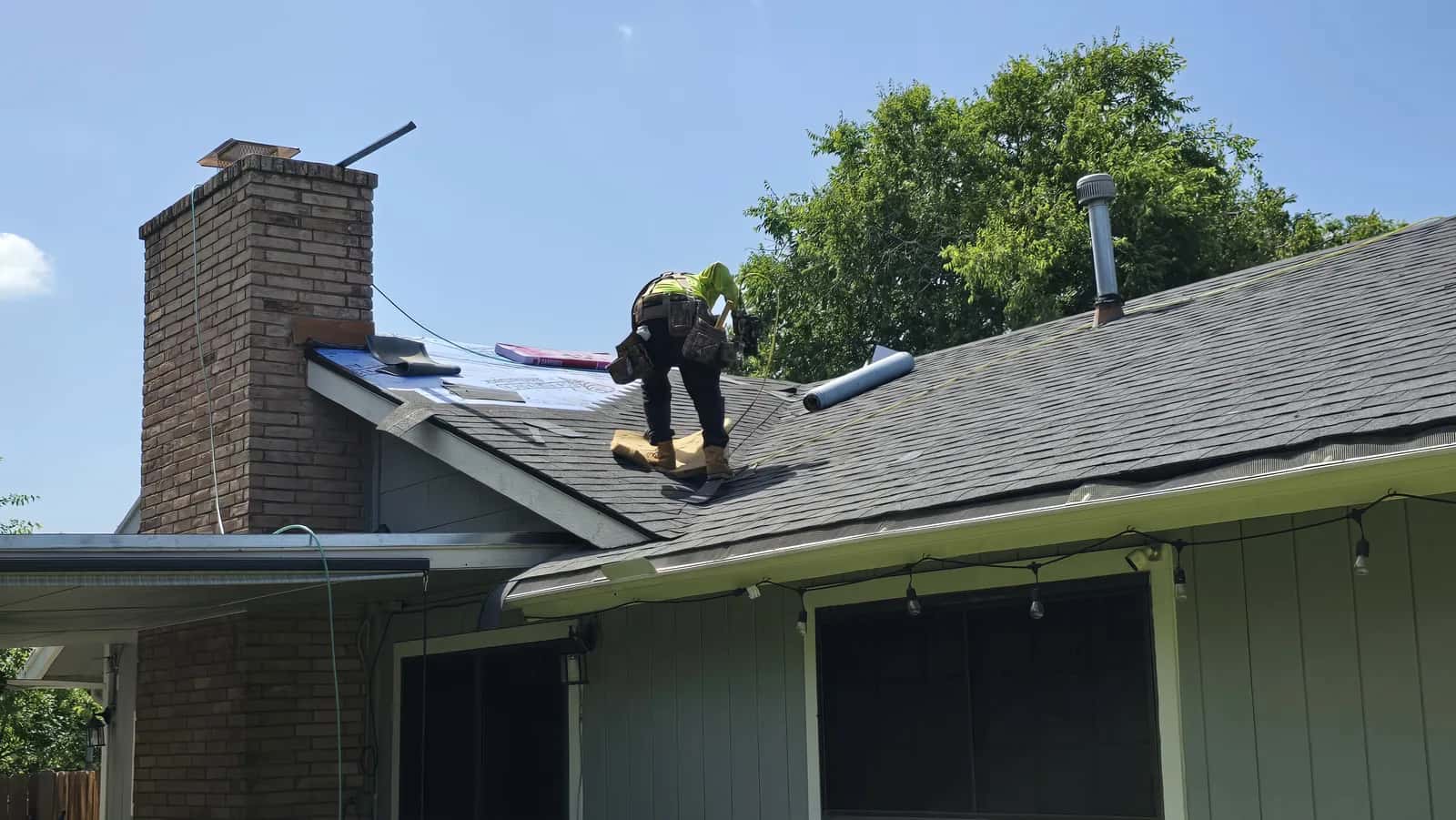 Roofer installing dark gray architectural shingles beside a brick chimney over blue synthetic underlayment