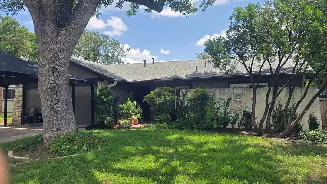 Single-story dark stone home with a completed gray shingle roof shaded by a large oak tree.