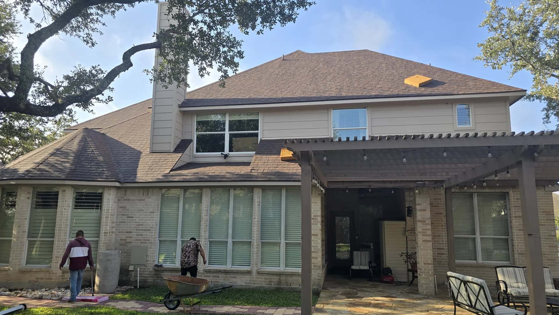 Rear of a two-story brick and stucco home with a finished brown architectural shingle roof, covered pergola patio and lawn.