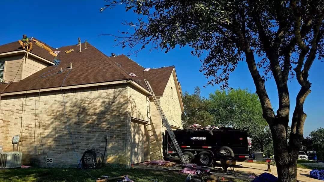 Rear of a two-story brick home with a new brown shingle roof going on, roofer at the ridge and a dump trailer beside the house.