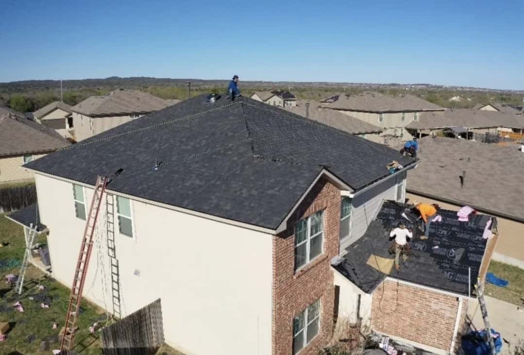 Elevated view of a two-story brick and stucco home with a crew installing dark shingles, ladder at the eave and underlayment on the lower roof.