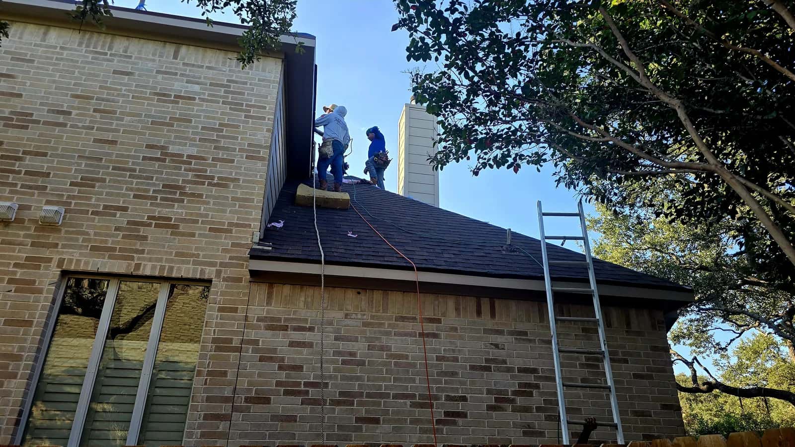 Roofers installing dark architectural shingles near the chimney of a two-story brick home, ladder set against the eave