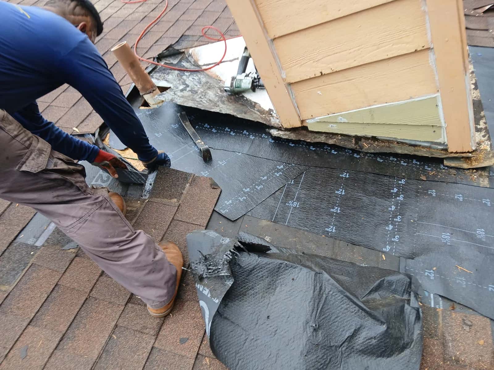 Roofer repairing flashing and decking where a brown shingle roof meets a sided wall, old felt pulled back