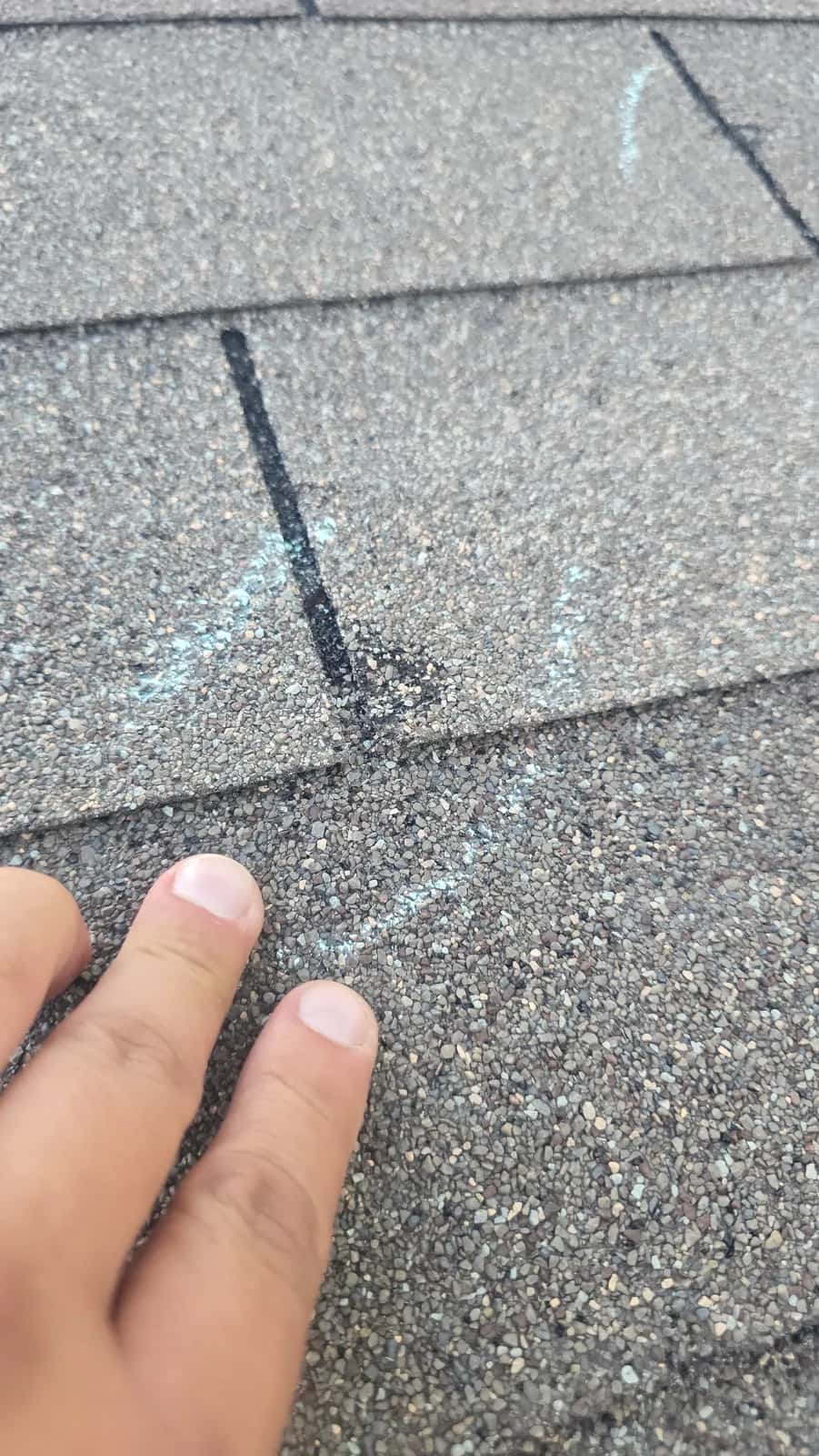 Close-up of a hand pointing to a chalk-circled hail impact on a gray asphalt shingle