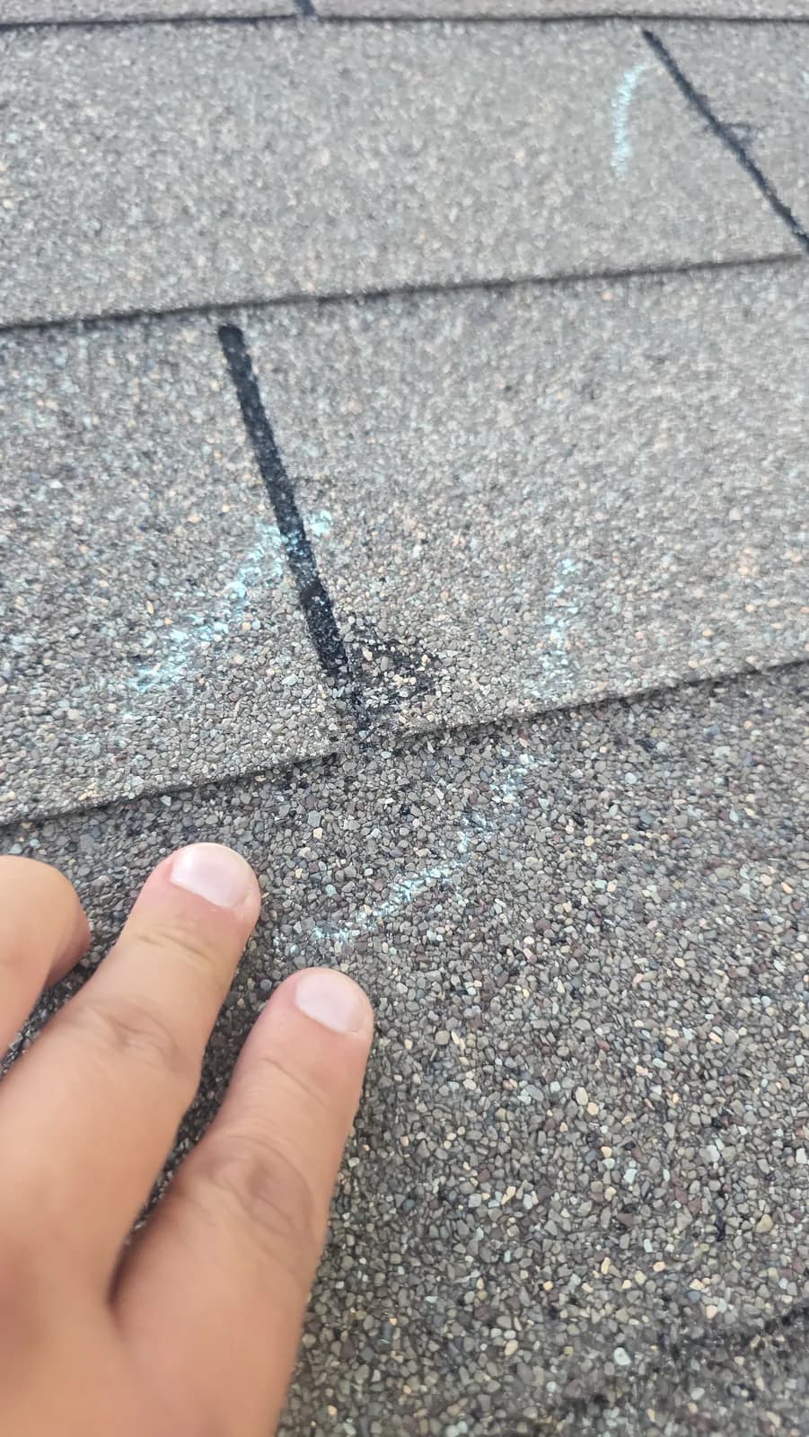 Close-up of a hand pointing to a chalk-circled hail impact on a gray asphalt shingle.