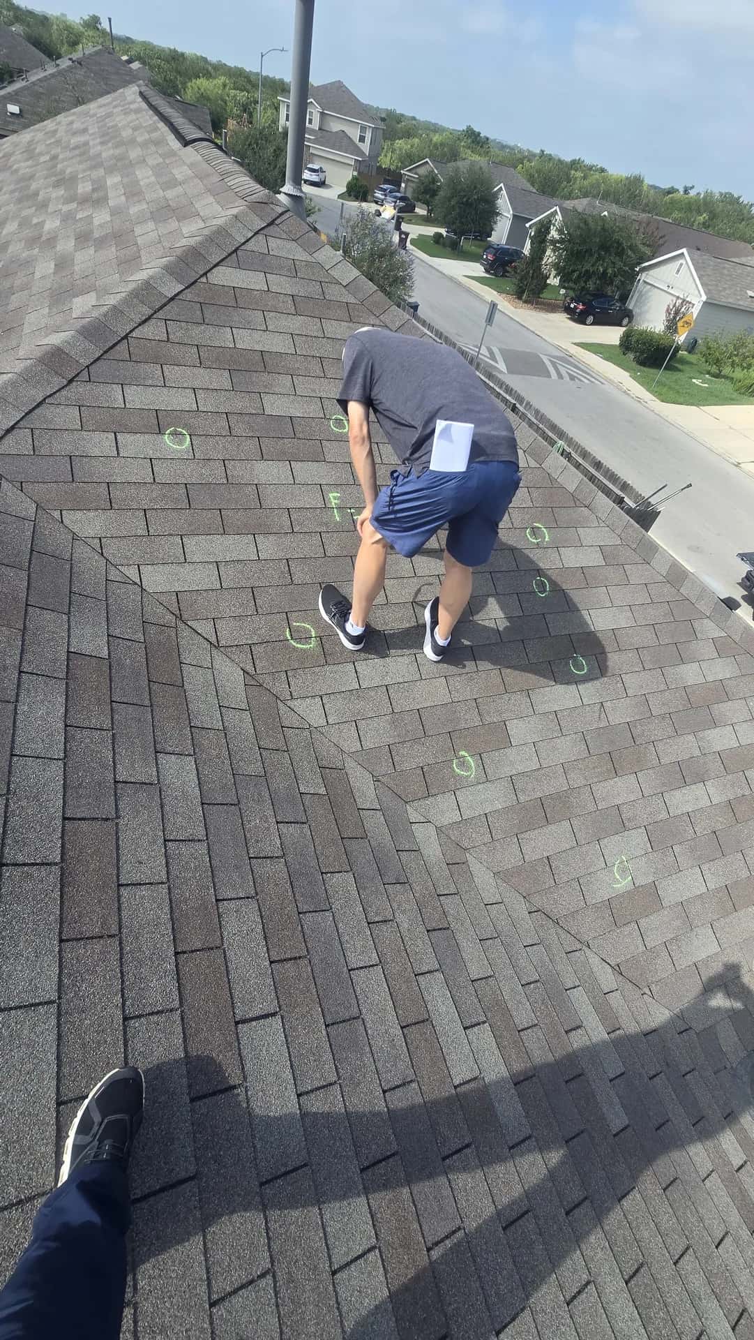 Inspector crouched on a brown shingle roof marking hail hits with chalk, residential street below
