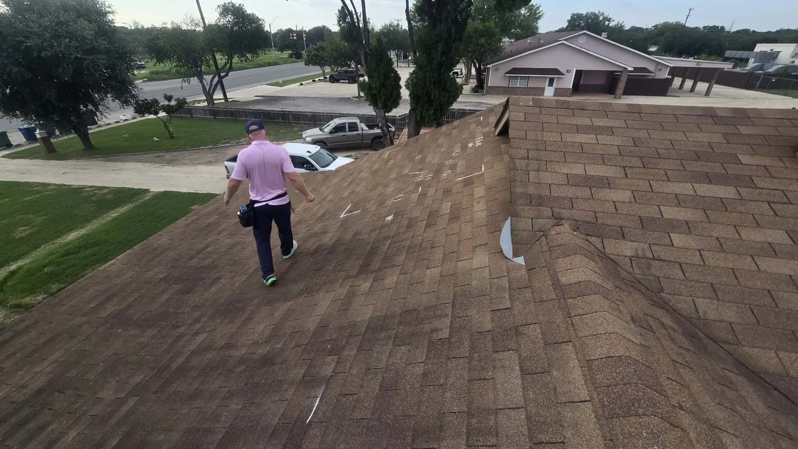 Inspector walking a brown shingle roof marked with chalk during a hail damage inspection, street and trees beyond.