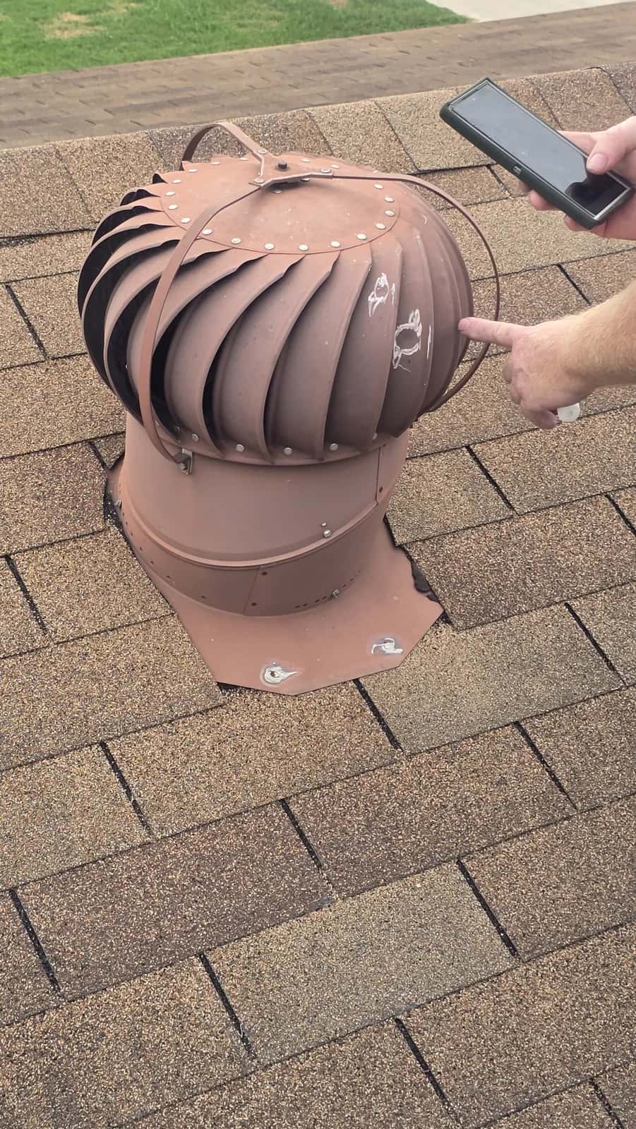Inspector pointing at a weathered, rust-stained turbine vent on a brown asphalt shingle roof during a roof inspection