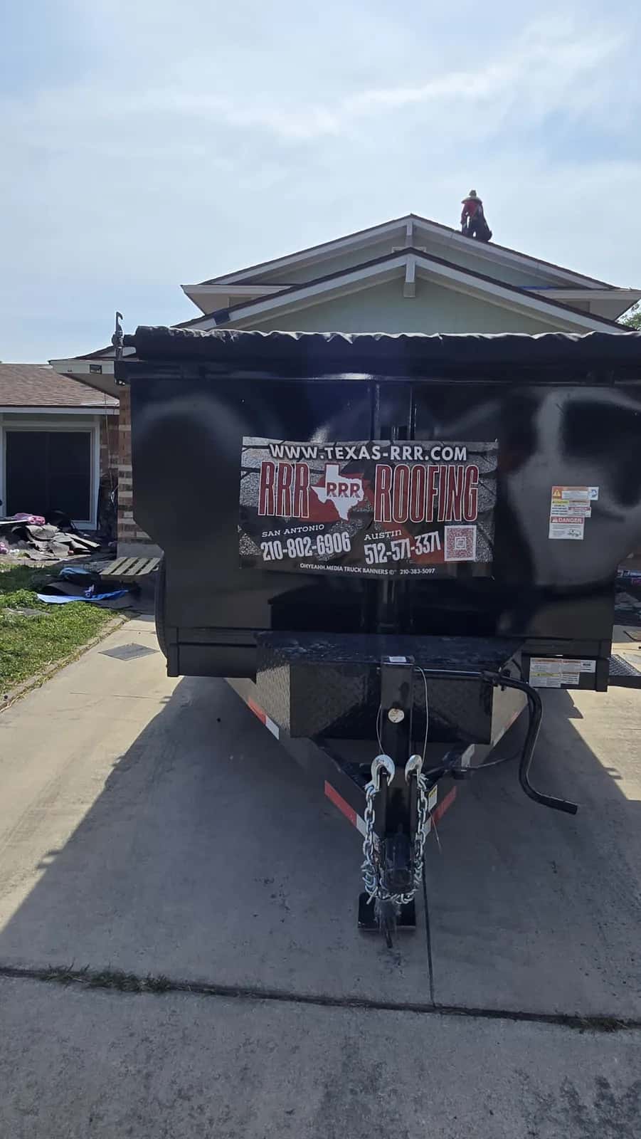 Black RRR Roofing branded dump trailer backed into a residential driveway with a crew member on the roof of the house behind it
